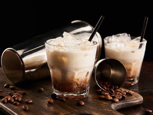 white russian cocktail in glasses with straws on wooden board with coffee grains and shaker isolated on black